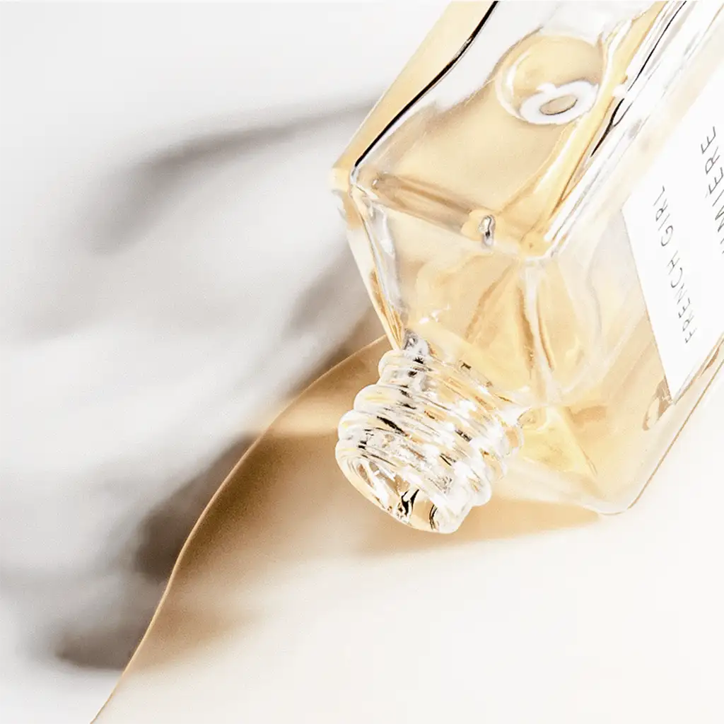 A clear glass perfume bottle with a gold-toned cap and label, containing a golden-hued liquid.