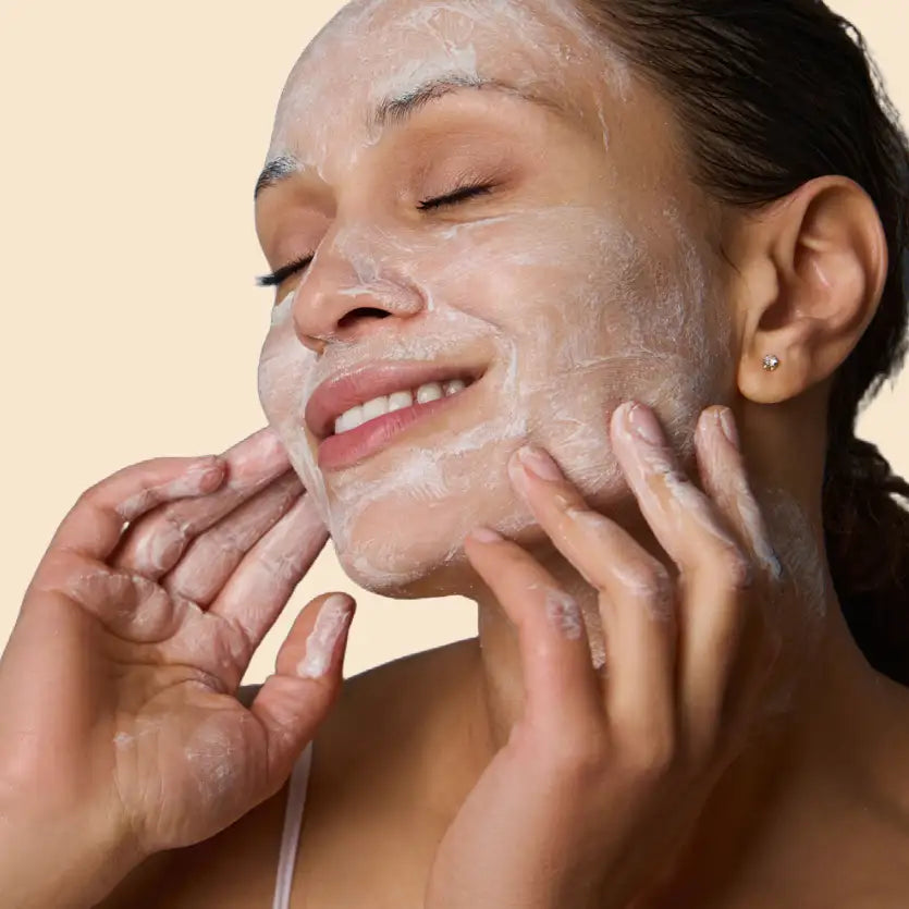 A woman with dark hair and a small diamond stud earring gently applies a thick, white facial mask to her face while smiling.